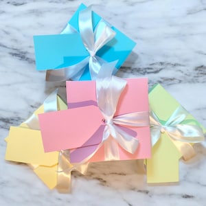 May include: A selection of pastel-coloured gift boxes in blue, pink, and yellow, each tied with a white satin ribbon. Each box has a matching blank tag attached, ready for personalisation. The boxes are arranged on a marble surface.