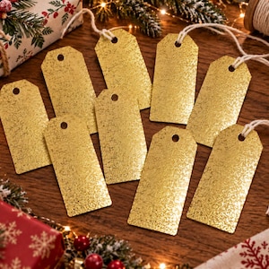 Könnte beinhalten: Ein Set aus acht rechteckigen goldenen Geschenkanhängern mit einer strukturierten, schimmernden Oberfläche. Jeder Anhänger hat oben ein kreisförmiges Loch und ist an einer weißen Schnur befestigt. Die Anhänger sind in einer Gruppe angeordnet.