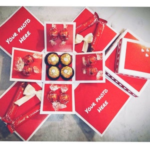 Pu&ograve; includere: Una scatola esplosiva rossa con bordi bianchi, piena di cioccolatini Lindt Lindor e Ferrero Rocher. La scatola ha spazi per le foto ed &egrave; decorata con fiocchi bianchi e barrette di cioccolato.