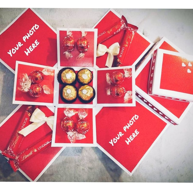 Pu&ograve; includere: Una scatola esplosiva rossa con bordi bianchi, piena di cioccolatini Lindt Lindor e Ferrero Rocher. La scatola ha spazi per le foto ed &egrave; decorata con fiocchi bianchi e barrette di cioccolato.