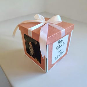 May include: A rose gold glitter gift box with a white ribbon tied in a bow. The box has a small framed picture on one side and a white label with the text "Happy Mothers day mum Lots of love".