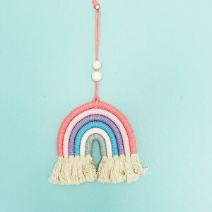 May include: A handmade macrame rainbow wall hanging. The rainbow features layers of pink, purple, blue, and gray yarn, with a fringe at the bottom. Two white beads and a pink string are at the top for hanging. The background is a light blue.