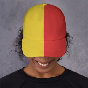 May include: A baseball cap with a split design, featuring a yellow panel on the left and a red panel on the right. The cap is worn by a person with dark curly hair and a black shirt. The background is a solid gray.