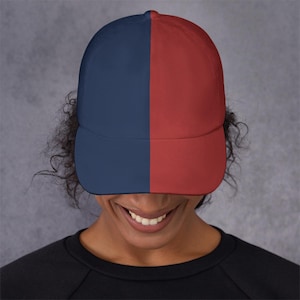 May include: A baseball cap with a split design, featuring a navy blue panel on the left and a red panel on the right. The cap has a curved brim and is made of a smooth fabric. The hat is worn by a person.