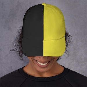 May include: A baseball cap with a split design, featuring a black panel on the left and a yellow panel on the right. The cap has a curved brim and a button on top. The hat is made of a soft material.