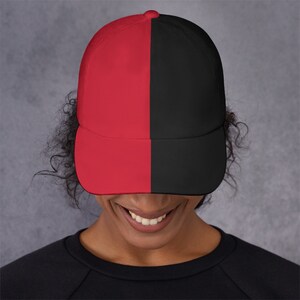Puede incluir: Una gorra de béisbol roja y negra con una división vertical en el medio. La gorra la lleva puesta una persona con el pelo rizado.