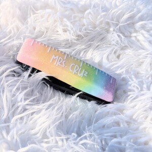 May include: A rainbow glitter stapler with a ruler on the top and the text "Mrs. Cruz" written on it.