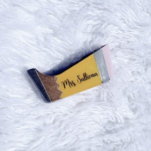 Puede incluir: Un porta lápices de color amarillo, marrón y plateado con purpurina con el texto "Mrs. Sullivan" en el lateral.