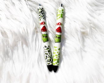 May include: Two green and white pens with a Grinch design and the word "HO HO" printed on them. The pens have a black leopard print design on one side and a green and white design on the other side.