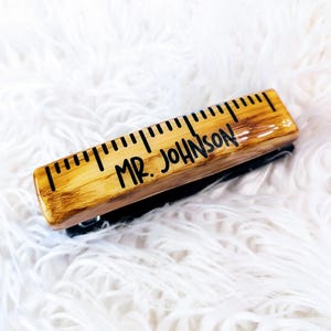 May include: A wooden ruler stapler with a black metal base. The ruler is marked with inches and has the name "MR. JOHNSON" written on it.