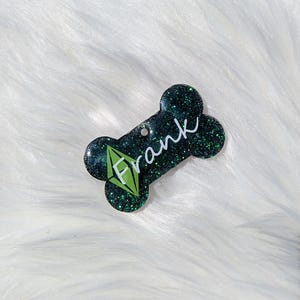 Puede incluir: Hueso de perro de purpurina verde con un diamante verde y el nombre "Frank" escrito en blanco.