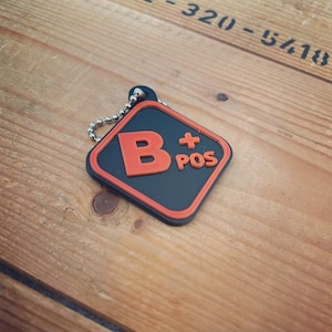 Könnte beinhalten: Ein schwarzer und orangefarbener quadratischer Schlüsselanhänger mit einer silbernen Kette. Der Schlüsselanhänger trägt die Buchstaben "B+ POS" aufgedruckt.