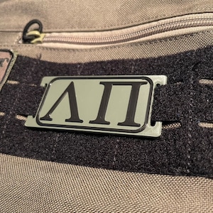 Könnte beinhalten: Ein silberner Metallaufnäher mit schwarzer Beschriftung, die "ΛΠΙ" lautet, befestigt an einem schwarzen und grauen Stoffband.