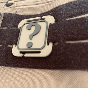 Puede incluir: Un parche de color beige con un signo de interrogación negro sobre una correa de velcro negra.