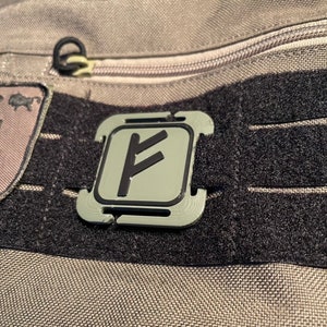 Peut inclure: Un sac en tissu gris et noir avec une poche zippée et une sangle en velcro noir. La sangle a une boucle en plastique grise avec un symbole noir dessus. Le symbole est une rune stylisée.