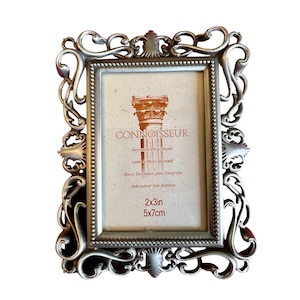 May include: Ornate silver-colored photo frame with scrolling details. The frame has a rectangular opening and includes the text "CONNOISSEUR" and "2x3in". The frame's dimensions are 2 inches by 3 inches, or 5 cm by 7 cm.