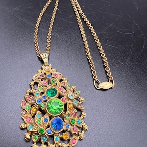 May include: Gold-toned pendant necklace with a floral design. The pendant features an array of colorful gemstones in shades of green, blue, pink, and yellow. The necklace has a delicate chain and a secure clasp.