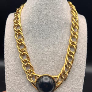 May include: A gold-toned chain necklace with a large, round, black center stone. The necklace is displayed on a gray mannequin. The chain links are thick and closely linked, creating a bold, statement piece of jewelry.