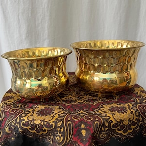 May include: Two gold metal planter pots with a hammered texture. The pots are sitting on a patterned fabric surface.