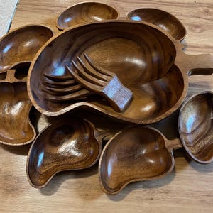 May include: A set of wooden serving bowls in a variety of shapes and sizes. The bowls are made of dark brown wood and have a smooth, polished finish. The set includes a large, oval-shaped bowl with a handle, as well as several smaller bowls in the shape of peppers. A wooden serving fork is included.