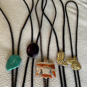 May include: Collection of bolo ties with various pendant designs. Includes a turquoise stone, an oval purple stone, a Native American thunderbird, a gold cowboy boot with rhinestones, and a gold horse head.