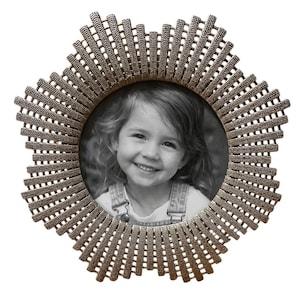 May include: A round, bronze-colored photo frame with a sunburst design. The frame has a textured, woven pattern radiating outwards from the circular opening. Inside the frame is a black and white photograph of a smiling child.