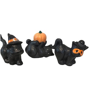 May include: Three black cat figurines. The first wears a black witch hat with an orange band. The second lies on its back holding an orange pumpkin. The third wears a black top hat and an orange eye mask. Each cat has textured black fur.