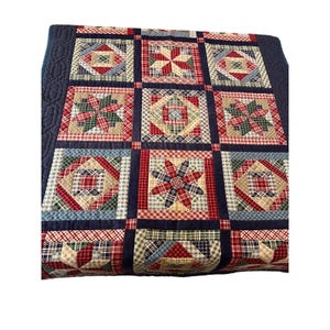 May include: A patchwork quilt with a navy blue border and a grid of squares. Each square features a different geometric pattern in red, blue, green, and beige plaid fabrics. The quilt is a traditional style.
