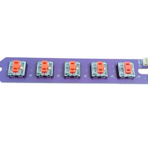 May include: A purple circuit board with five red and clear mechanical keyboard switches. The board has a small notch on the left side and a row of pins on the right side.