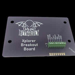 May include: A black Xplorer Breakout Board with the RetroCult logo and text. It features a green terminal block and gold-coloured pins. The board has text labels for various functions like HOME, UP, LEFT, RIGHT, SEL, START, and GND.