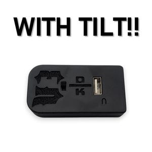 以下が含まれることがあります： 上部に「WITH TILT!!」というテキストが記載された黒い長方形の電子機器。この機器にはUSBポート、小さなスイッチ、および上面にDとKの文字を含む浮き彫りの刻印があり、左端は丸みを帯びた形状をしています。
