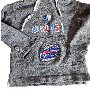 Puede incluir: Sudadera gris con capucha, cuello alto y bolsillo delantero. La sudadera presenta el texto "Go Bills" en letras coloridas y un parche con el logotipo de los Buffalo Bills. El cordón es blanco con un diseño estampado.