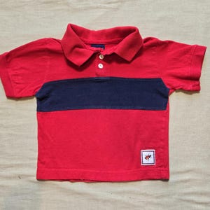 May include: A short-sleeved polo shirt in bright red with a wide horizontal navy blue stripe across the chest. The shirt features a collared neckline with two white buttons and a small square patch on the lower right side depicting a beetle.