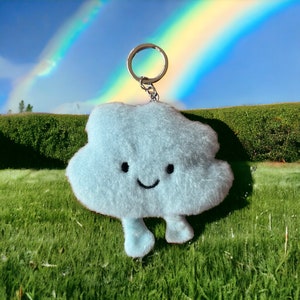May include: A light blue plush cloud keychain with a smiling face and two small legs. The keychain is hanging from a silver key ring and is in front of a green grassy field with a rainbow in the background.