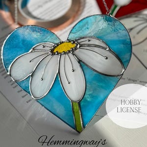 Puede incluir: Un patrón de vitral de una margarita blanca en un corazón azul. El patrón se titula "Hemingway's Flower Heart Stained Glass Pattern".