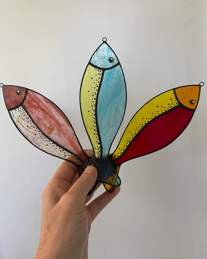Peut inclure: Mobile de poissons en vitrail color&eacute; avec trois poissons stylis&eacute;s en rouge, jaune et bleu. Chaque poisson a un contour noir et une petite boucle pour l'accrocher. Les poissons sont maintenus ensemble &agrave; la base.