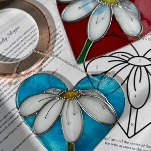 May include: Two stained glass heart-shaped ornaments with a white daisy design. The first ornament has a red background and the second ornament has a blue background.