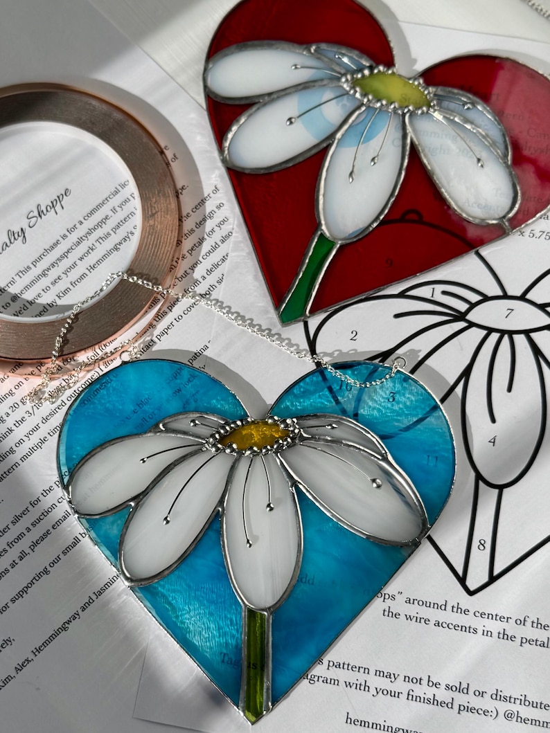 May include: Two stained glass heart-shaped ornaments with a white daisy design. The first ornament has a red background and the second ornament has a blue background.