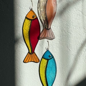 Peut inclure: Trois poissons d&eacute;coratifs en vitrail color&eacute;s suspendus devant un mur blanc textur&eacute;. Les poissons sont dans des tons d'orange, de rouge, de jaune, de rose et de bleu. Chaque poisson a un crochet en m&eacute;tal pour l'accrochage.