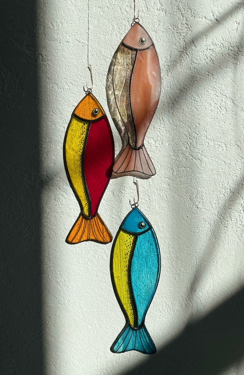 Peut inclure: Trois poissons d&eacute;coratifs en vitrail color&eacute;s suspendus devant un mur blanc textur&eacute;. Les poissons sont dans des tons d'orange, de rouge, de jaune, de rose et de bleu. Chaque poisson a un crochet en m&eacute;tal pour l'accrochage.