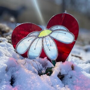May include: A red glass heart with a white daisy flower in the center. The heart and flower are on a bed of snow.