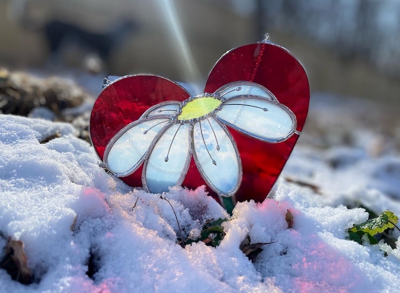 May include: A red glass heart with a white daisy flower in the center. The heart and flower are on a bed of snow.