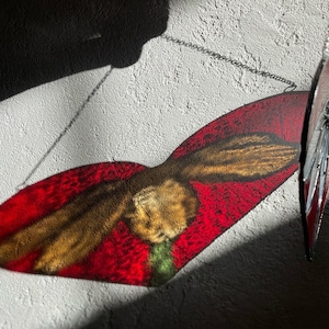 May include: A red and gold stained glass hummingbird hanging from a silver chain against a white textured wall.