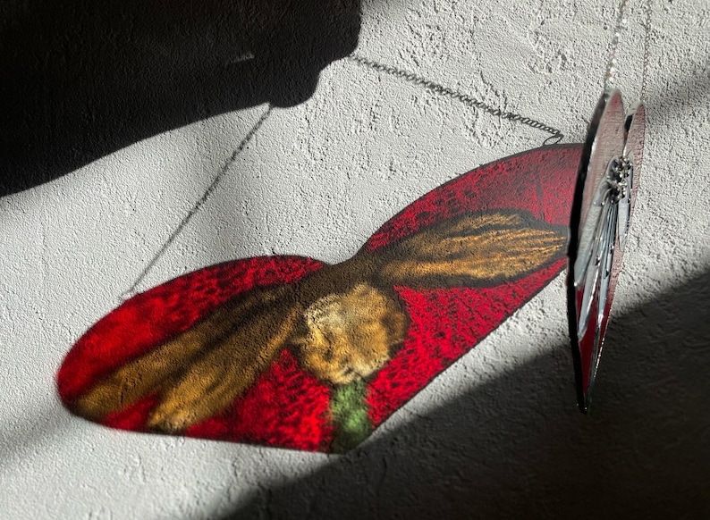 May include: A red and gold stained glass hummingbird hanging from a silver chain against a white textured wall.