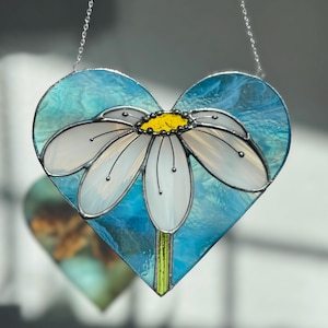 May include: A stained glass heart-shaped ornament with a white daisy flower on a blue background. The ornament is hanging from a silver chain.