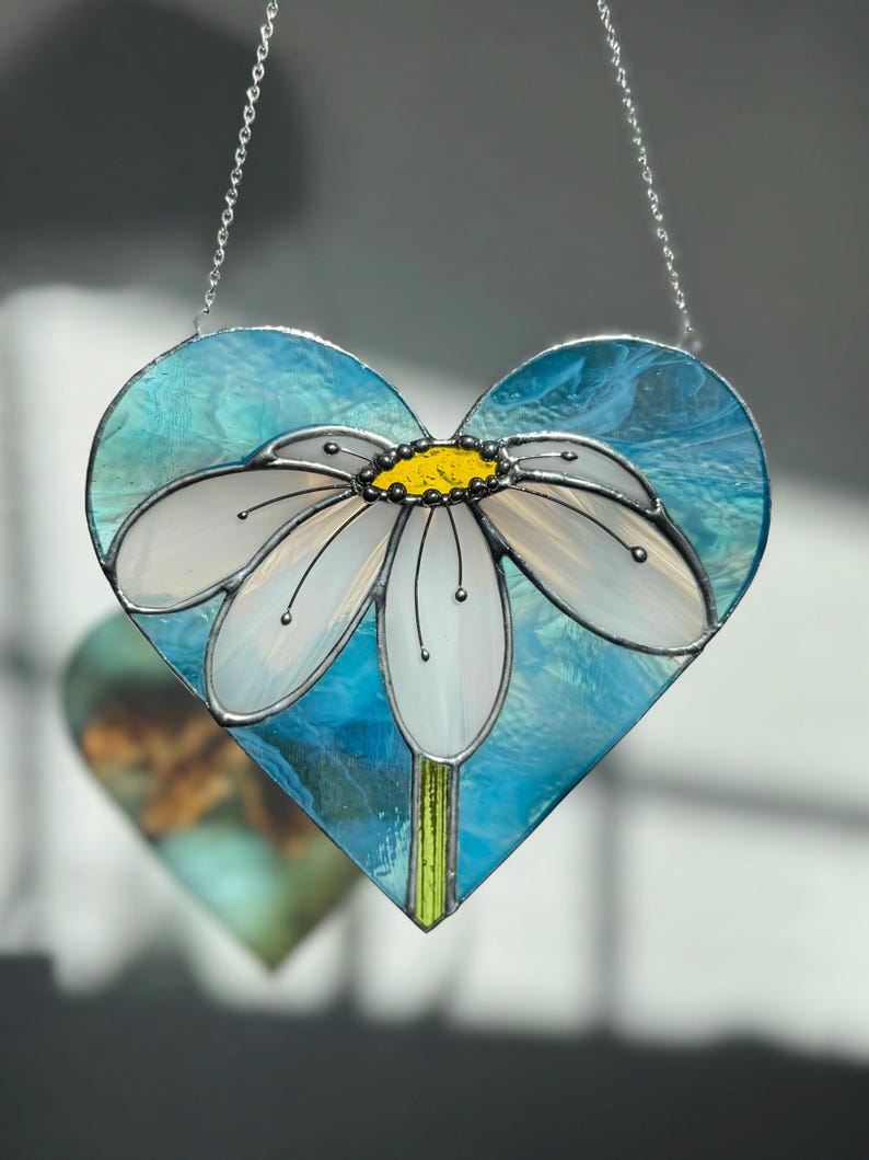 May include: A stained glass heart-shaped ornament with a white daisy flower on a blue background. The ornament is hanging from a silver chain.