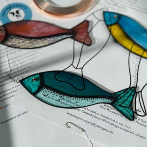 Peut inclure: Gros plan sur un poisson en vitrail dans les tons de turquoise, de rouge et de bleu, avec des d&eacute;tails noirs. Le poisson est sur une surface blanche avec du texte et un crochet. Le poisson mesure 16.19 cm H x 5.4 cm L.