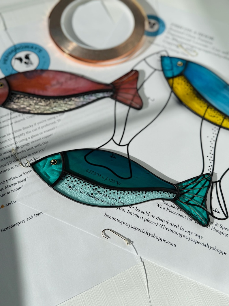 Peut inclure: Gros plan sur un poisson en vitrail dans les tons de turquoise, de rouge et de bleu, avec des d&eacute;tails noirs. Le poisson est sur une surface blanche avec du texte et un crochet. Le poisson mesure 16.19 cm H x 5.4 cm L.