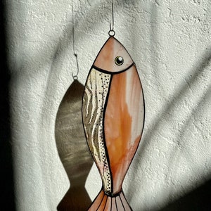 Peut inclure: Un attrape-soleil en forme de poisson en vitrail, aux tons p&ecirc;che et cr&egrave;me. Il pr&eacute;sente un motif textur&eacute; sur un c&ocirc;t&eacute; et un &oelig;il clair. Suspendu &agrave; un fil fin, il projette une ombre sur le mur.