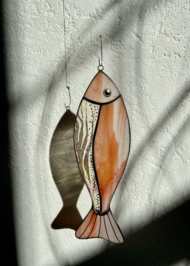 Peut inclure: Un attrape-soleil en forme de poisson en vitrail, aux tons p&ecirc;che et cr&egrave;me. Il pr&eacute;sente un motif textur&eacute; sur un c&ocirc;t&eacute; et un &oelig;il clair. Suspendu &agrave; un fil fin, il projette une ombre sur le mur.